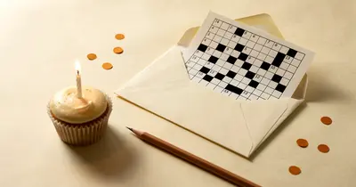 Un cruciverba stampato infilato nella busta di un biglietto d’auguri, accanto a un cupcake con una candela accesa, su carta color crema.